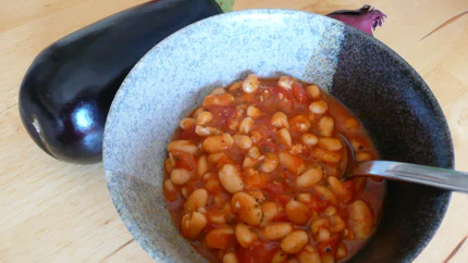 Weiße Bohnen in Tomatensauce mit Pfeffer und Kräutern in einer Schale. Darum liegen Zutaten.