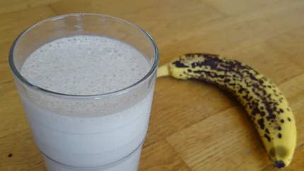 Ein Glas schaumiger Bananenmlich steht auf dem Tisch, daneben liegt eine reife Banane.