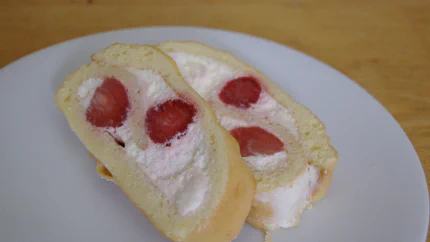 Zwei Scheiben einer mit Schlagsahne und Erdbeeren gefüllten Biskuitroulade liegen auf einem Teller. Die eine lehnt an der zweiten.