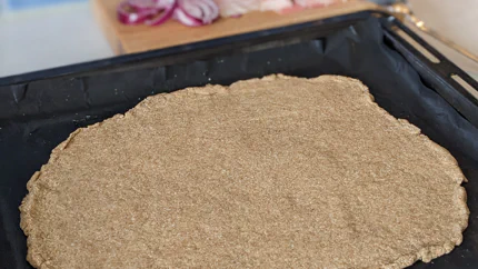 Auf einem Backblech liegt ein hellbrauner Teig aus Goldleinsamen. Darum liegen Zutaten für Pizza.