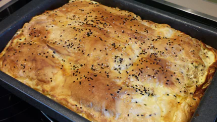 In einer Kastenform liegt ein Börek mit Spinat und Feta. Die oberste Schicht des Teigs ist braun gebacken und Sesam und Schwarzkümmel bestreut.