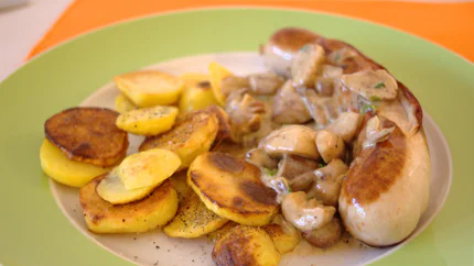 Eine von beiden Seiten braun gebratene Kalbsbratwust. Daneben liegt eine Portion Bratkartoffeln, dazwischen eine Sahnesauce mit Steinpilzen.