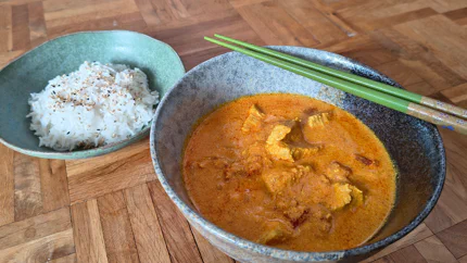 Eine Portion Butterhuhn aus groben Hühnerstücken und Tomaten-Joghurt-Sauce ist in einer Schale. Daneben steht eine zweite Schale mit gekochtem Reis.