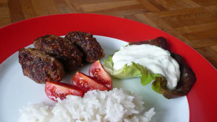 Drei Cevapcici liegen neben Reis, Salat und einem Joghurtdip auf einem bunten Teller.
