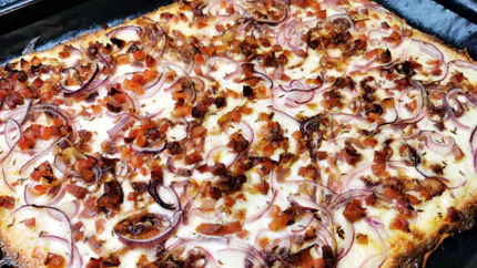 Ein Blech mit Flammkuchn mit braunem Speck und roten Zwiebelringen.