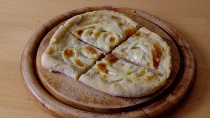 Ein ovaler Flammkuchn mit hohem Rand und Zwiebelringen. Er hat einzelne, braune Stellen.