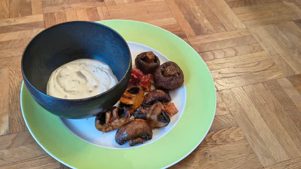 Braun gebackene und halbierte Champignons liegen auf einem Teller mit Joghurtdip mit Kräutern.