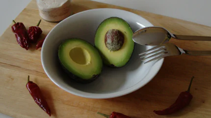 Eine halbierte Avocado liegt auf einem Holzbrett umgeben von Chilischoten und Salz.