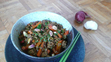 Eine Schale mit gebratenem Hackfleisch mit Paprika, Rüble, Petersilie und Pistazien. Dekoriert mit Essstäbchen, Zwiebel und Knoblauch.