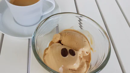 Zwei Kugeln Kaffeeeis liegen in einer Glasschale neben einem Espresso. Das Eis ist mit zwei Kaffeebohnen verziert.