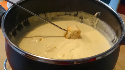 Ein Topf mit geschmolzenem Käse und eine Fonduegabel, auf der ein Brotwürfel aufgespießt ist.