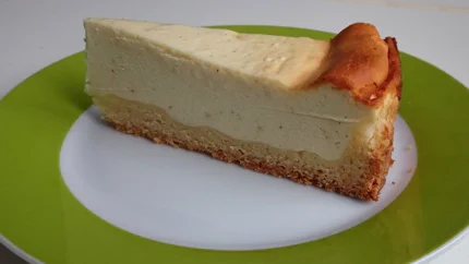 Ein Stück Käsekuchen auf einem Teller. Es hat einen Boden und ist in der Mitte cremig zart und am Rand leicht braun.