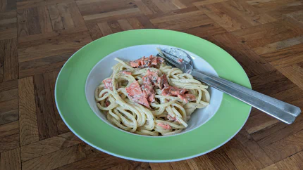 Ein tiefer Teller mit Spaghetti, Sahnesauce und Lachsscheiben. Es liegen Gabel und Löffel im Teller.