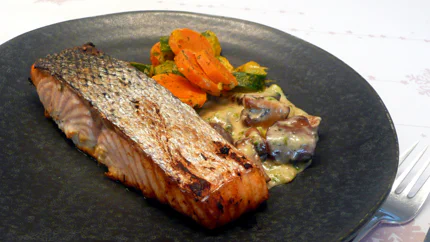 Ein Lachsfilet mit knuspriger Haut liegt neben Gemüse und Pilzen auf einem Teller.