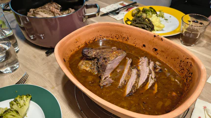 Ein Römertopf mit einer angeschnittenen Lammkeule in Bratensauce steht auf einem gut gedeckten Tisch.
