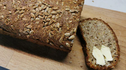 Ein Laib Brot mit Sonnenblumenkernen auf einem Holzbrett. Eine Scheibe wurde abgeschnitten, die mit Butter bestrichen ist.