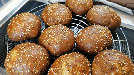 Neun dunkelbraune Brötchen liegen auf einem Rost. Auf ihnen sind Sesam und Kümmelkörner eingebacken.