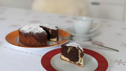 Ein Stück Marmorkuchen mit Puderzucker steht vor dem Rest des Kuchens. Eine Gabel und Tasse Kaffee stehen daneben.