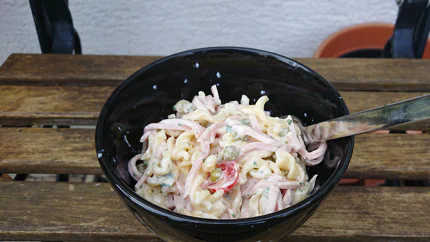 Nudelsalat mit Lyoner, Joghurt, Tomaten und Erbsen in einer Schale mit einer Gabel.