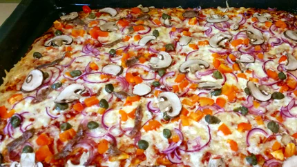 Eine Pizza auf einem Blech mit viel Mozzarella, Champignonscheiben, Kapern und Paprika.