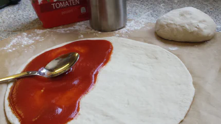 Ein ausgewallter Hefeteig auf einem Stück Backpapier. Eine Hälfte ist bereits mit Tomatensauce bestrichen.