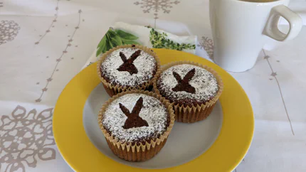 Drei dunkelbraune Muffins aus Rüben und Nüssen mit Puderzucker bestreut, der die Form eines Osterhasenkopfes hat. Dahinter steht eine Tasse Kaffee.