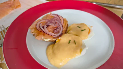 Ein halbiertes Ei übergossen mit gelblicher Sauce Hollandaise liegt neben einem Lachsbrötchen mit Zwiebelringen auf einem Teller.