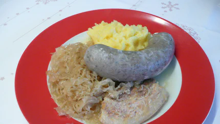 Eine Portion Sauerkraut und Kartoffelbrei auf einem Teller. Dazu gibt es eine Scheibe Bauchspeck und eine grobe Leberwurst.