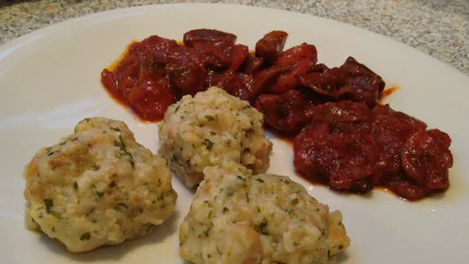 Drei Semmelknödel mit Kräutern liegen auf einem Teller vor einem dickflüssigen Ragout.
