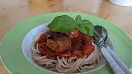 Eine Portion Spaghetti mit etwas Tomatensauce und drei Fleischbällchen darauf. Sie sind mit einer Spitze Basilikum garniert und es stecken Gabel und Löffel in den Spaghetti.
