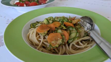 Eine Portion Spaghetti mit Zucchini- und Rübenscheiben in einem tiefen Teller, in dem auch Gabel und Löffel stecken. Dahinter steht ein Teller mit Tomaten-Mozzarella-Salat.