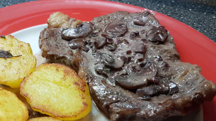 Ein gebratenes Steak mit Sauce und Pilzen neben goldbraunen Kartoffeln mit Rosmarinzweigen auf einem Teller.