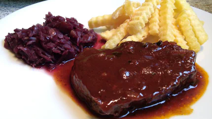 Eine Scheibe Tafelspitz unter einer dickflüssigen Bratensoße, dazu gibt es Rotkraut und geriffelte Pommes.