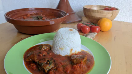 Grobe Rindfleischstücke liegen in einer dickflüssigen Sauce aus Tomaten und Gewürzen neben Reis in einem Teller. Dahinter steht ein Keramiktopf und einige Zutaten.