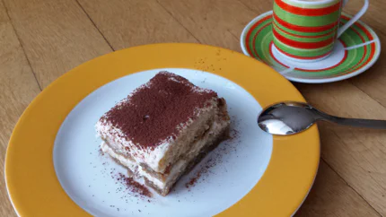 Eine Portion Tiramisu liegt auf einem Teller neben einem Espresso auf einem hölzernen Tisch.