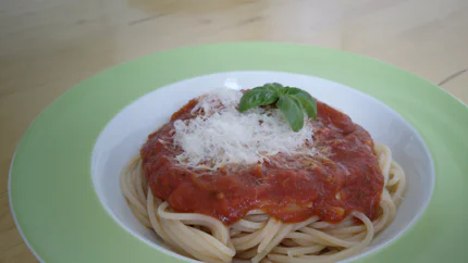 Eine Portion Spaghetti mit Tomatensauce bedeckt in einem tiefen Teller. Sie ist mit geriebenem Parmesan und einer Spitze Basilikum garniert.
