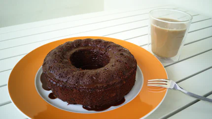 Ein brauner Kuchen aus einer Ringform mit groben Poren, an dem der Guss aus Zartbitterschokolade herunter läuft. Daneben steht eine Tasse Kaffee.