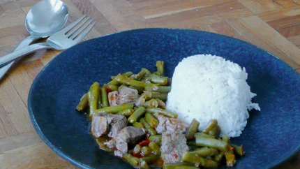 Eine Portion grüner Bohnen und Rindfleischstücken in einer würzigen Soße mit Reis in einem tiefen Teller auf einem Holztisch.