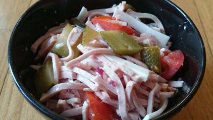 Eine Schale mit Wustsalat aus Lyoner, Gewürzgurken, Gruyère, Tomaten, Essig und Öl.