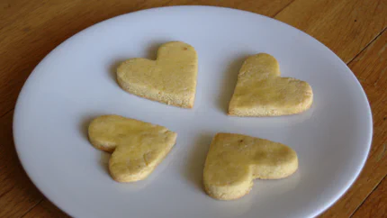 Vier goldbraune Zitronenplätzchen in Herzform liegen symmetrisch verteilt auf einem weißen, flachen Teller.