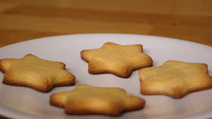 Vier Zitronenplätzchen in Ternform und dunkelbraunen Zacken liegen symmetrisch verteilt auf einem weißen Teller auf einem Holztisch.