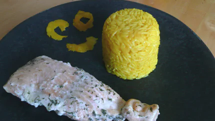 Eine Portion gelber Reis in Form gedrückt und auf einen Teller gestürzt. Daneben liegen Streifen einer Zitronenschale und ein Lachsfilet.