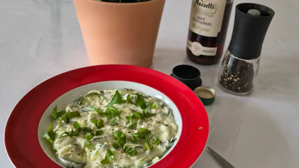 Eine Portino Zucchini-Nudeln mit Schafskäse-Sauce, Pfeffer und Basilikum in einem tiefen Teller mit breitem Rand. Daneben steht eine Flasche Rotweinessig.
